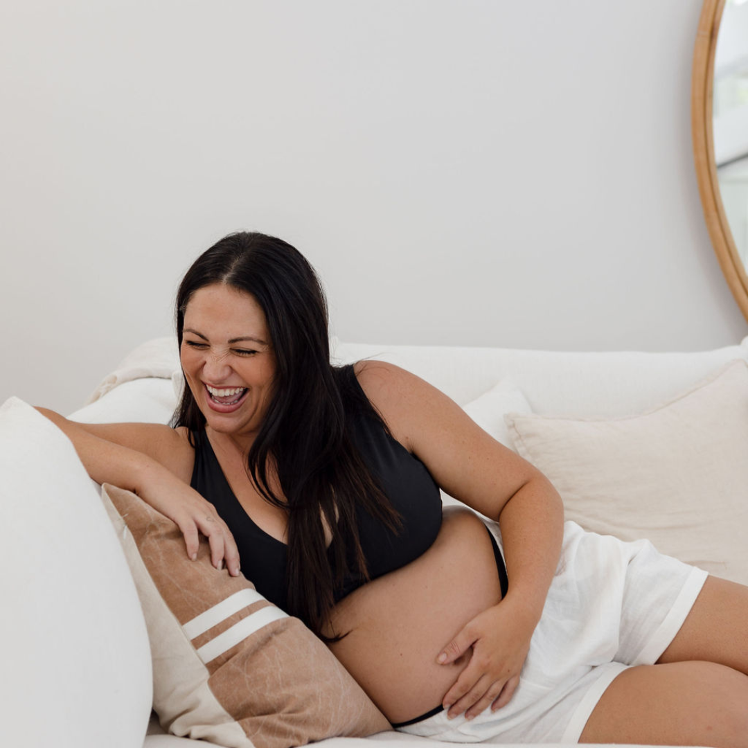 A mum-to-be reclines sideways on a white couch. She is holding her pregnant stomach and laughing, excited about pregnancy. 