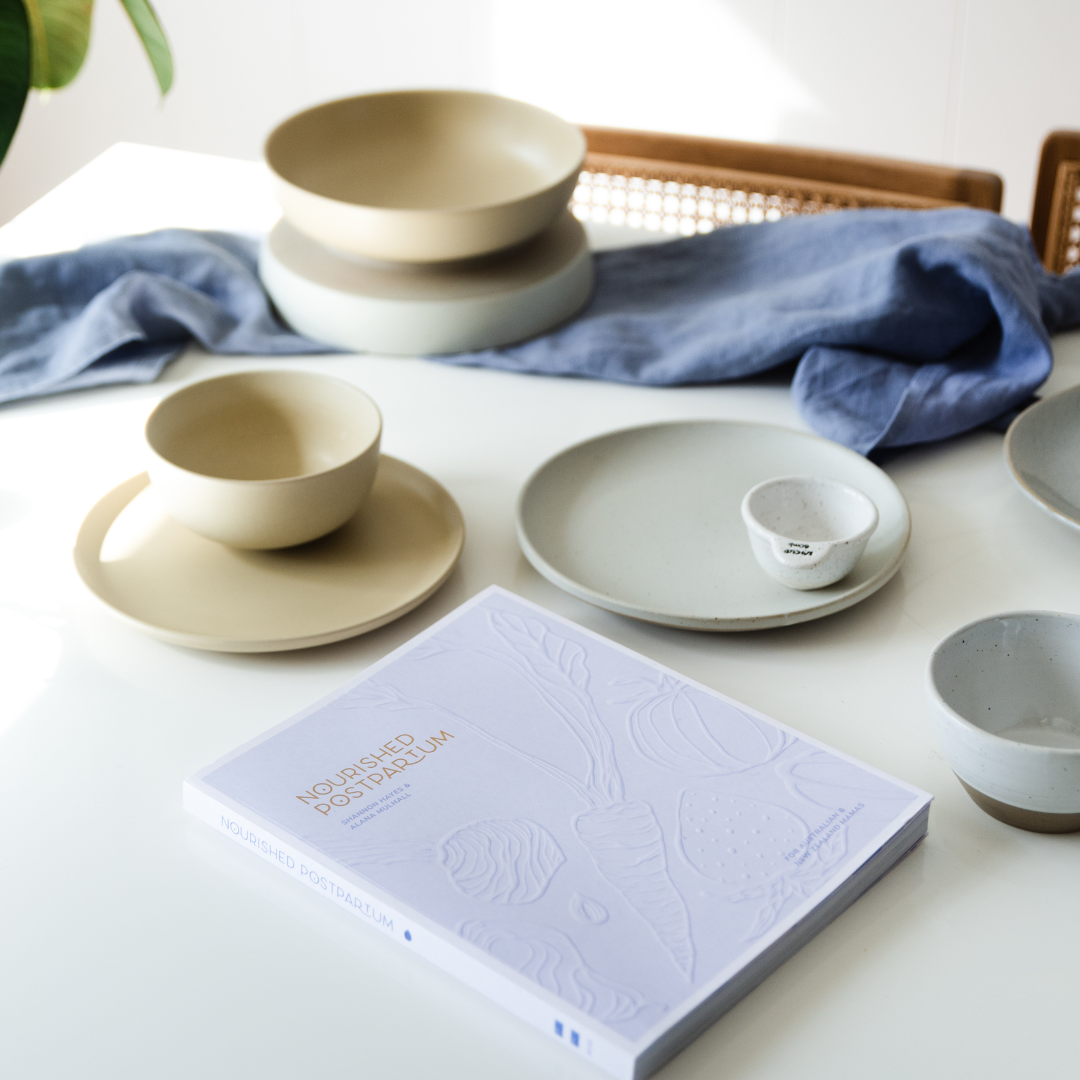 A table with Nourished Postpartum plates, bowls, and a book by Mothers Mylk on it.
