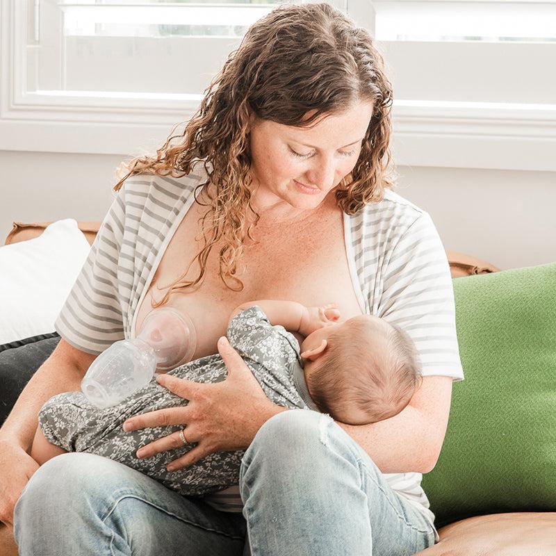 A woman breastfeeds her baby and is using the Haakaa Silicone Breast Pump 100ml Gen 1 by Haakaa on a couch.