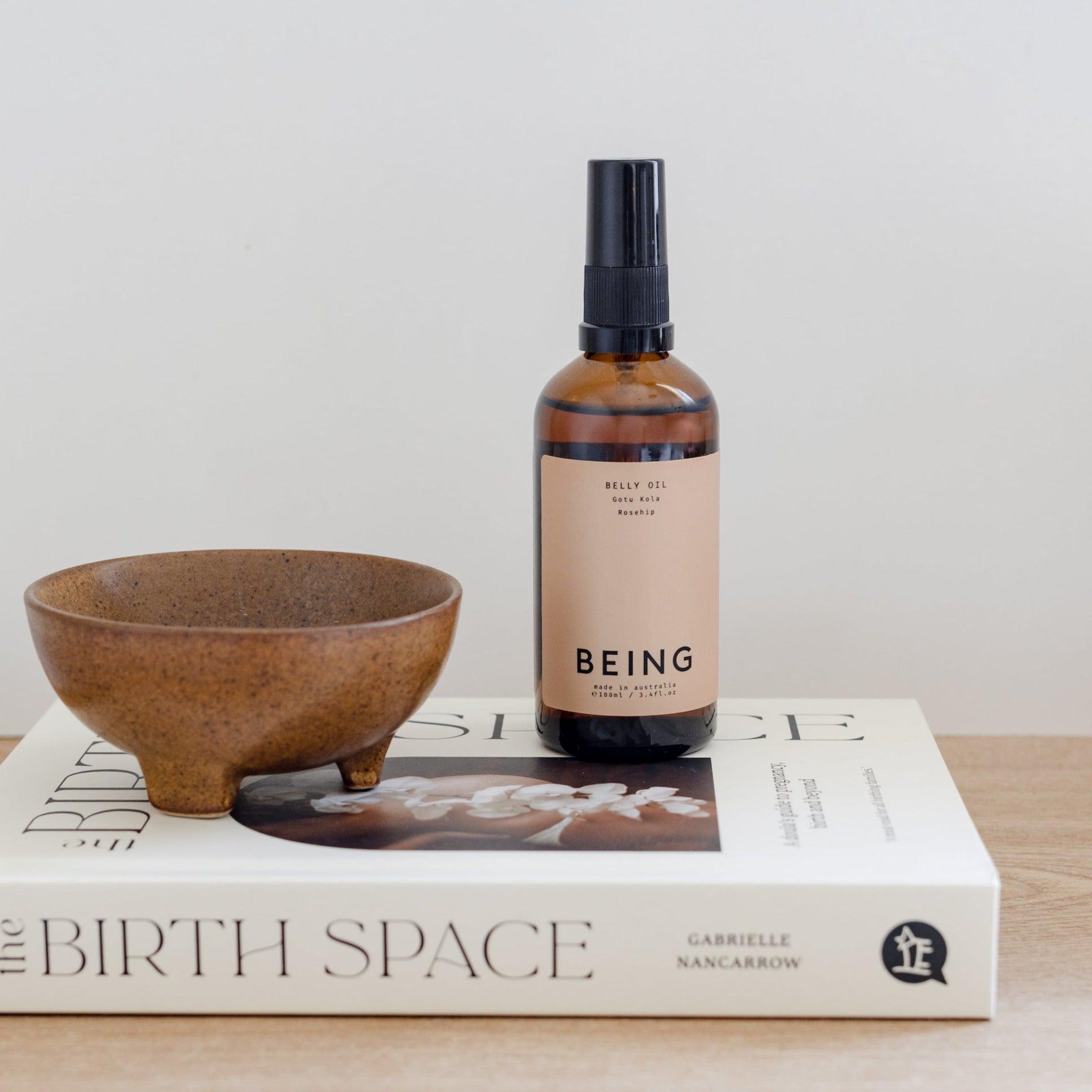 A book, a bowl, and a bottle of Belly Oil by Being Skincare on a table.