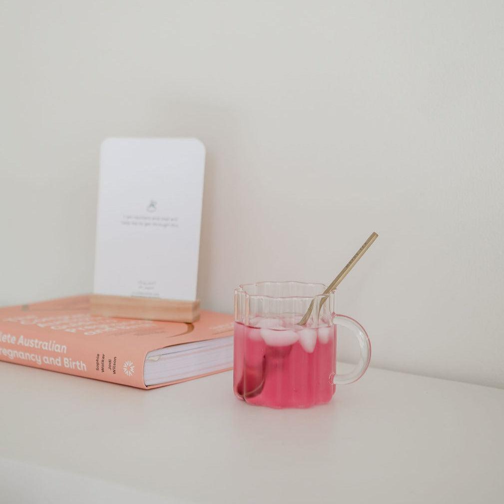 A cup of  Mini+Me. Hydramama Watermelon + Lime next to a book on a shelf.
