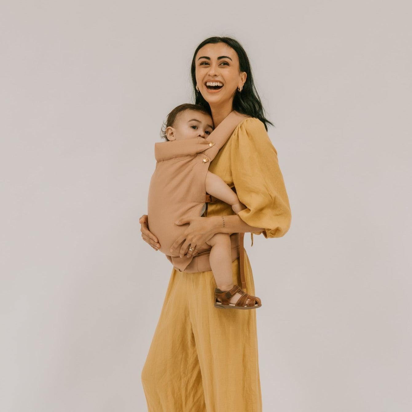 A woman wearing a Cinta Clip Carrier by Chekoh holds a baby in a yellow jumpsuit.