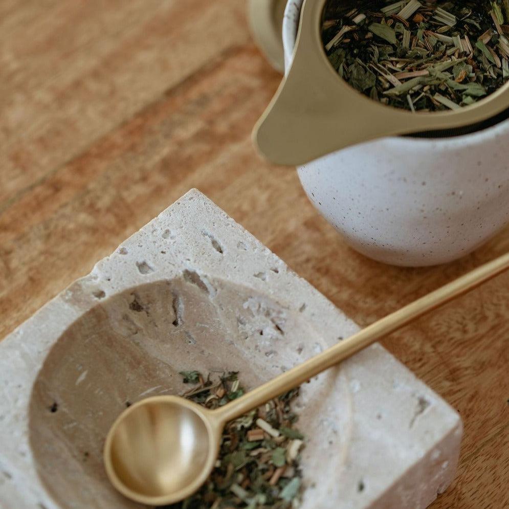 A pot of Mammae bosom ritual infusion nursing tea and a spoon, promoting breastfeeding, displayed on a wooden table.