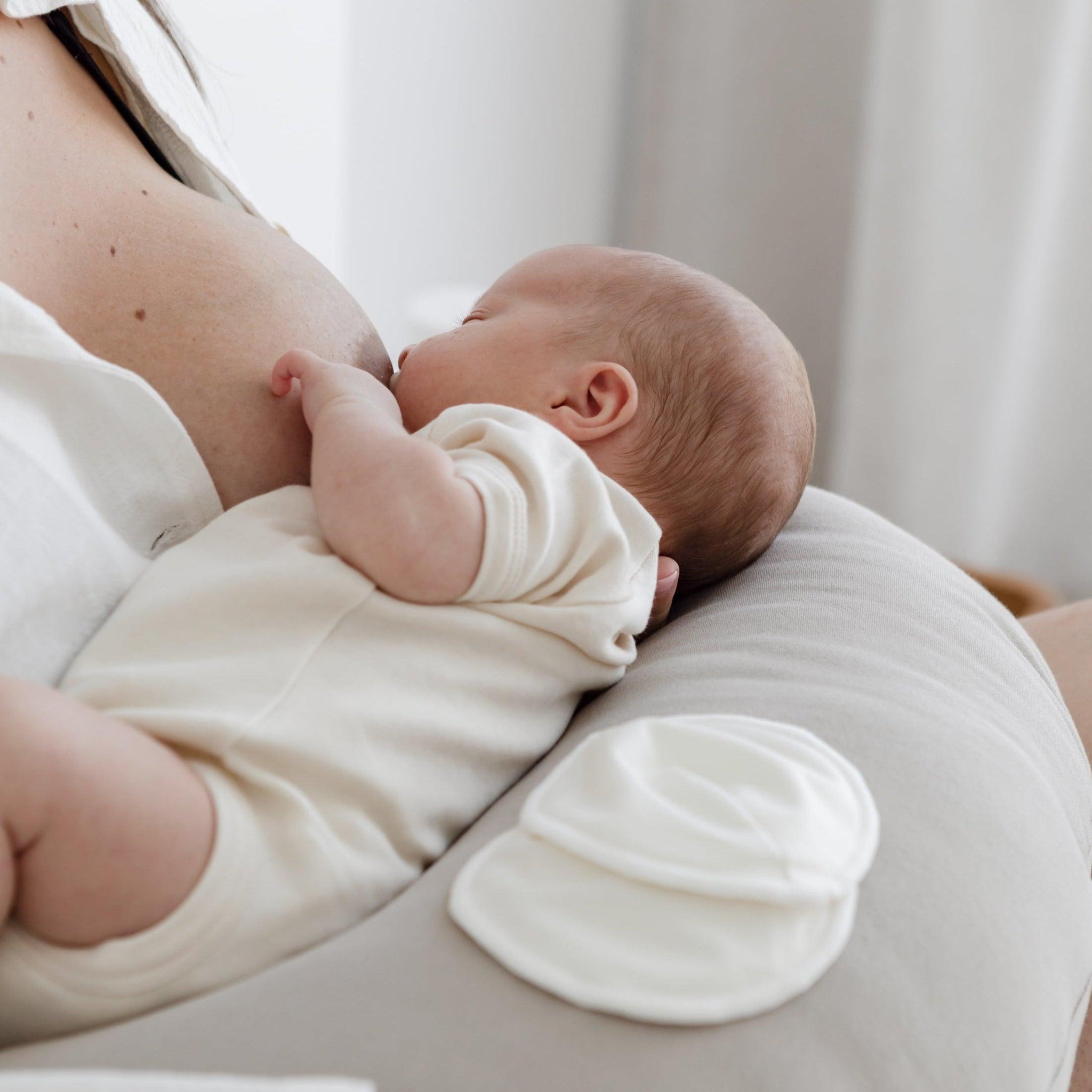 A woman is breastfeeding a baby in a white Mammae shirt, using bosom wearables for breast care during breastfeeding.