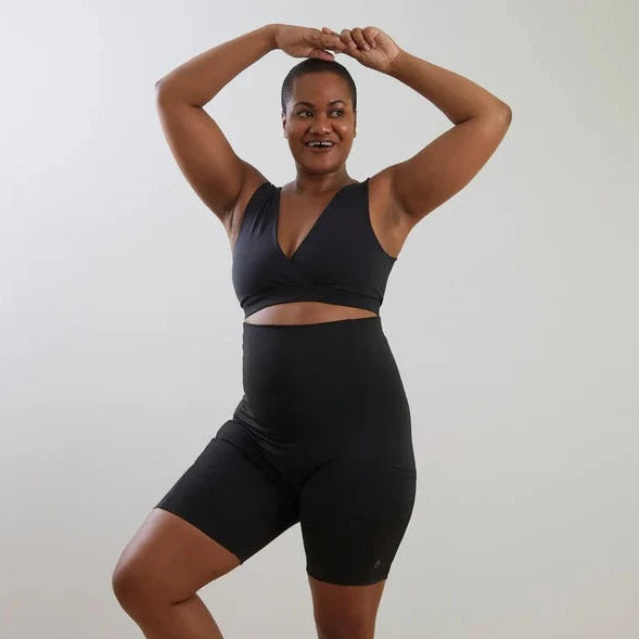 A woman in Bare Mum postpartum recovery shorts poses with her hands above her head and one knee lifted, standing against a white background.