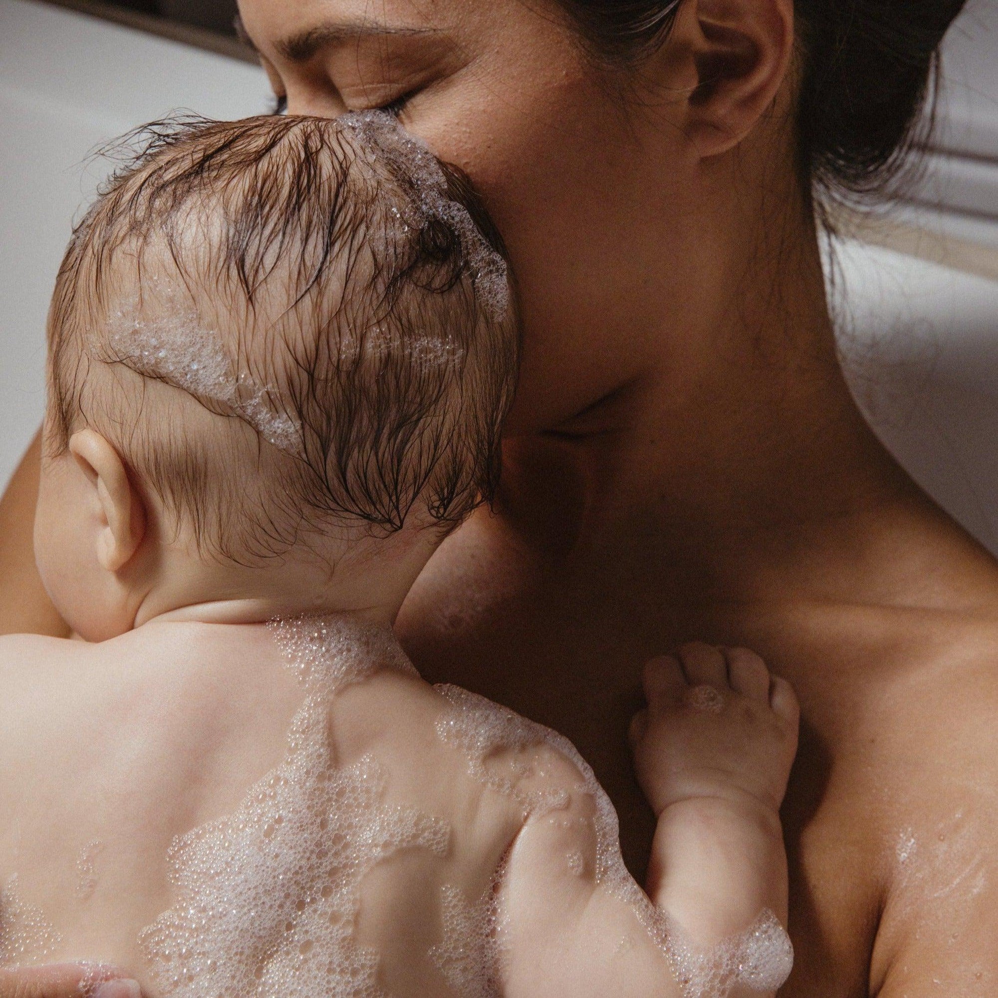 A woman is holding a baby in a bath tub filled with Wiley Body organic aloe vera-infused Hand & Body Wash.
