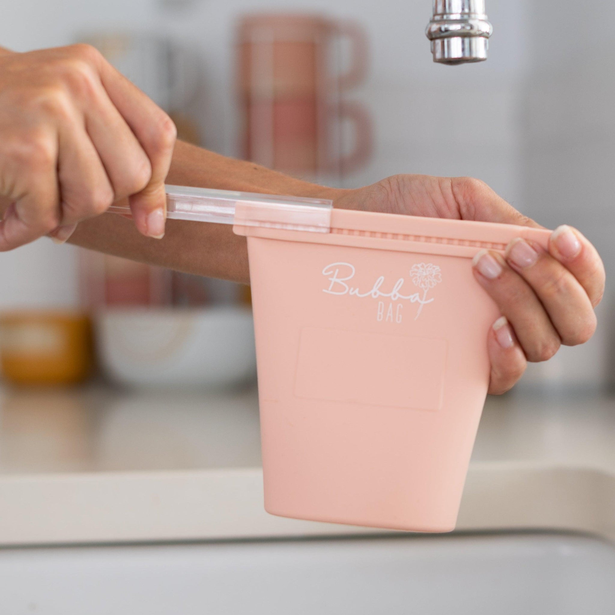 A person is holding a Marigold Baby pink plastic cup in front of a sink.