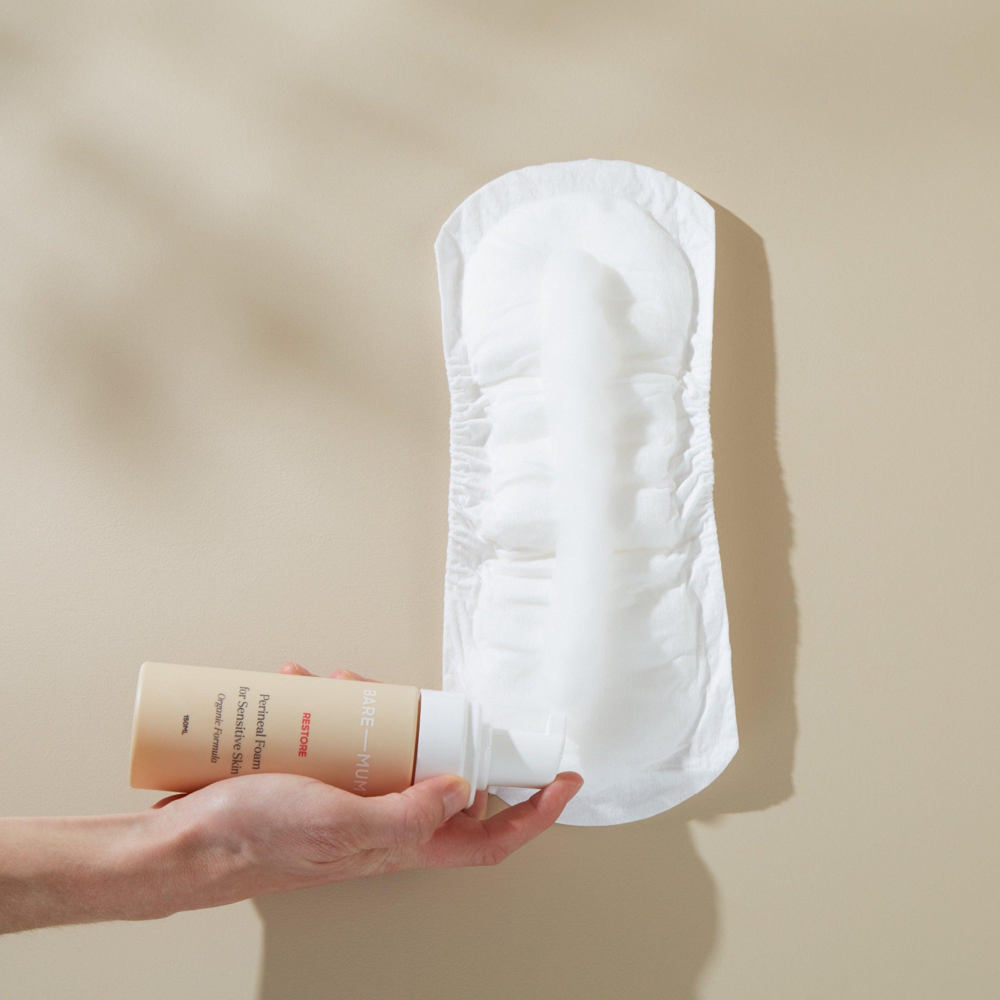 A hand holding a bottle of Bare Mum Perineal Foam and a Postpartum Pad for postpartum recovery on a beige background.