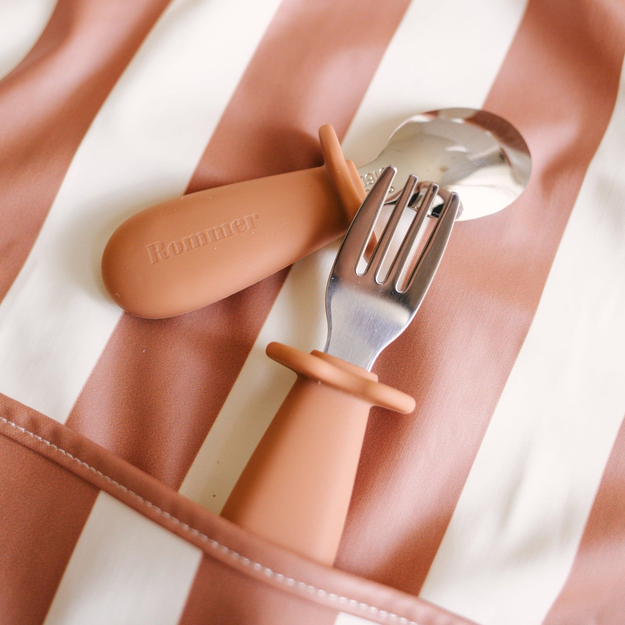 A Rommer toddler cutlery set in cinnamon, designed to encourage independence during mealtime for curious little hands, neatly stored in a striped bag.
