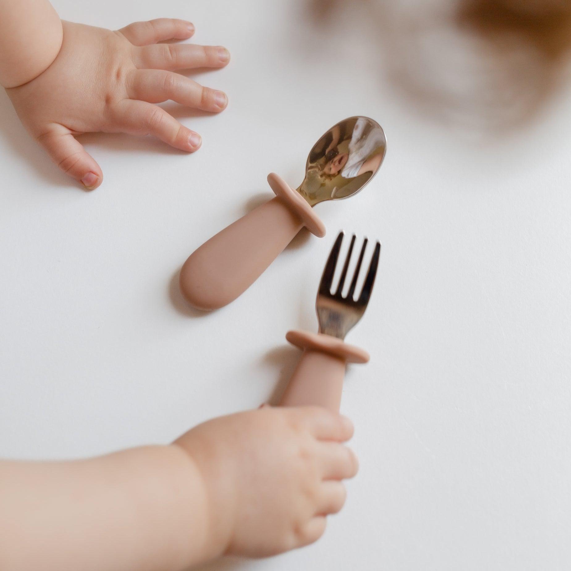 A Rommer toddler cutlery set in nude, being held by a toddler's hand during mealtime.