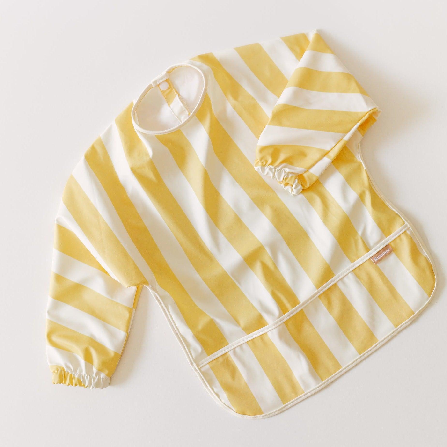 The Rommer Smock Bib, made from super soft fabric, features yellow and white striped colour variations on a white surface.