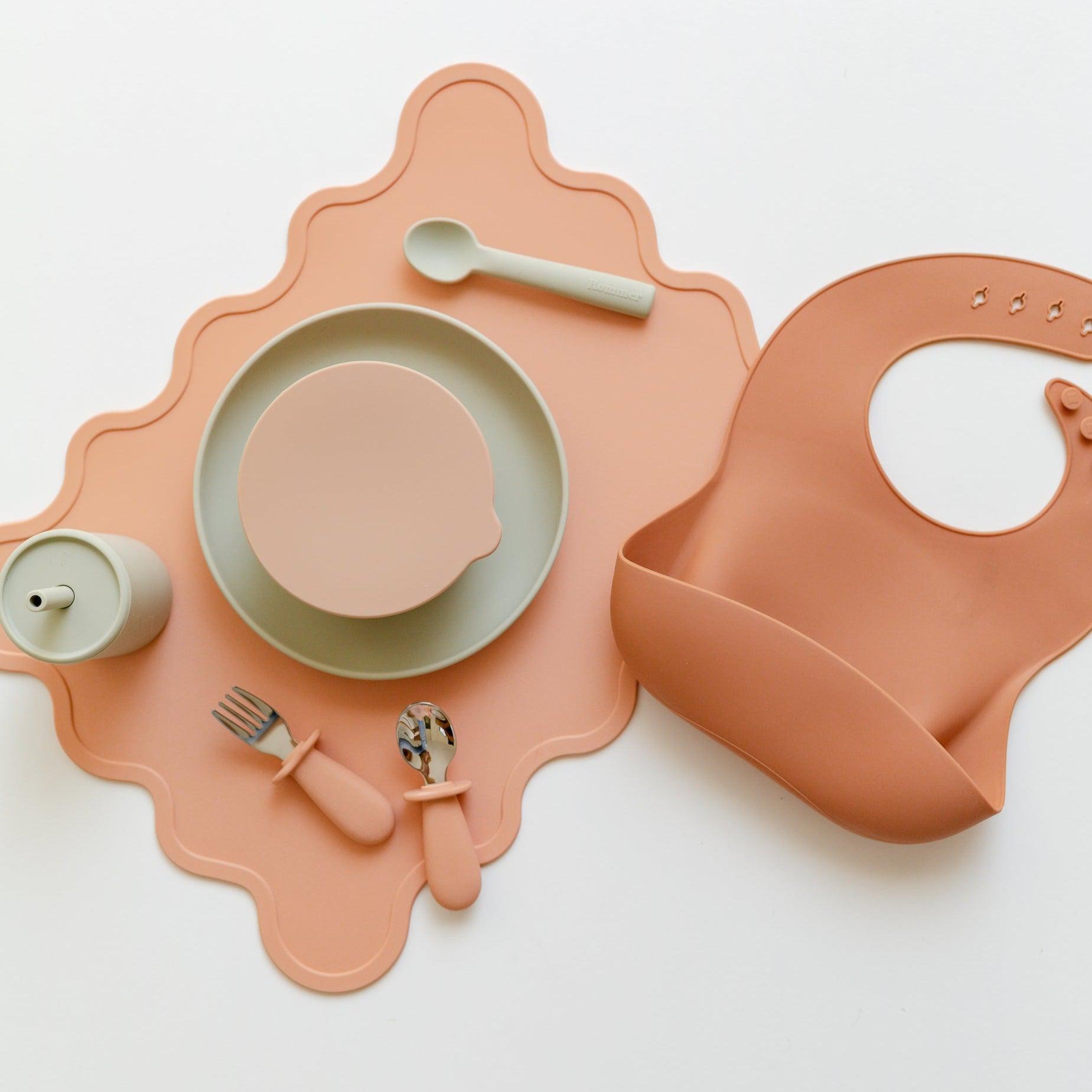 A Rommer toddler cutlery set in melon, comprising stainless steel utensils and a silicone bib.