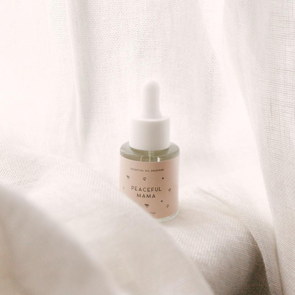 A small bottle of Peaceful Mama Essential Oil Dropper from Seasons of Mama sitting on top of a white sheet.