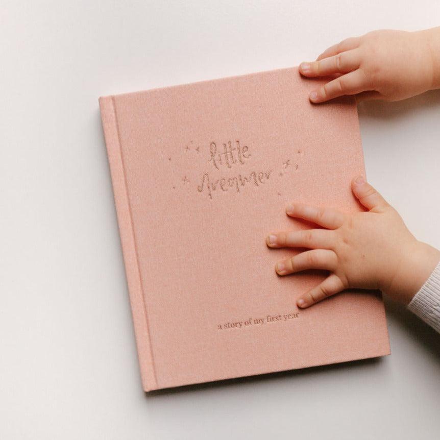A baby holding a Emma Kate Co Little Dreamer Baby Journal in Petal.