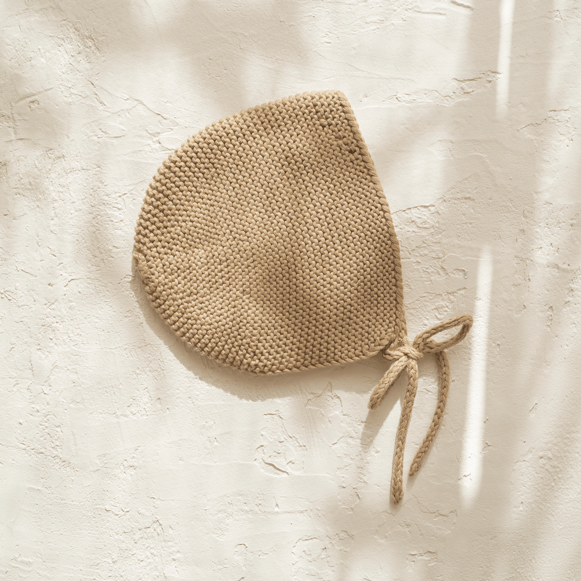 An illoura baby bonnet | olive, made of organic cotton, hanging on a wall.