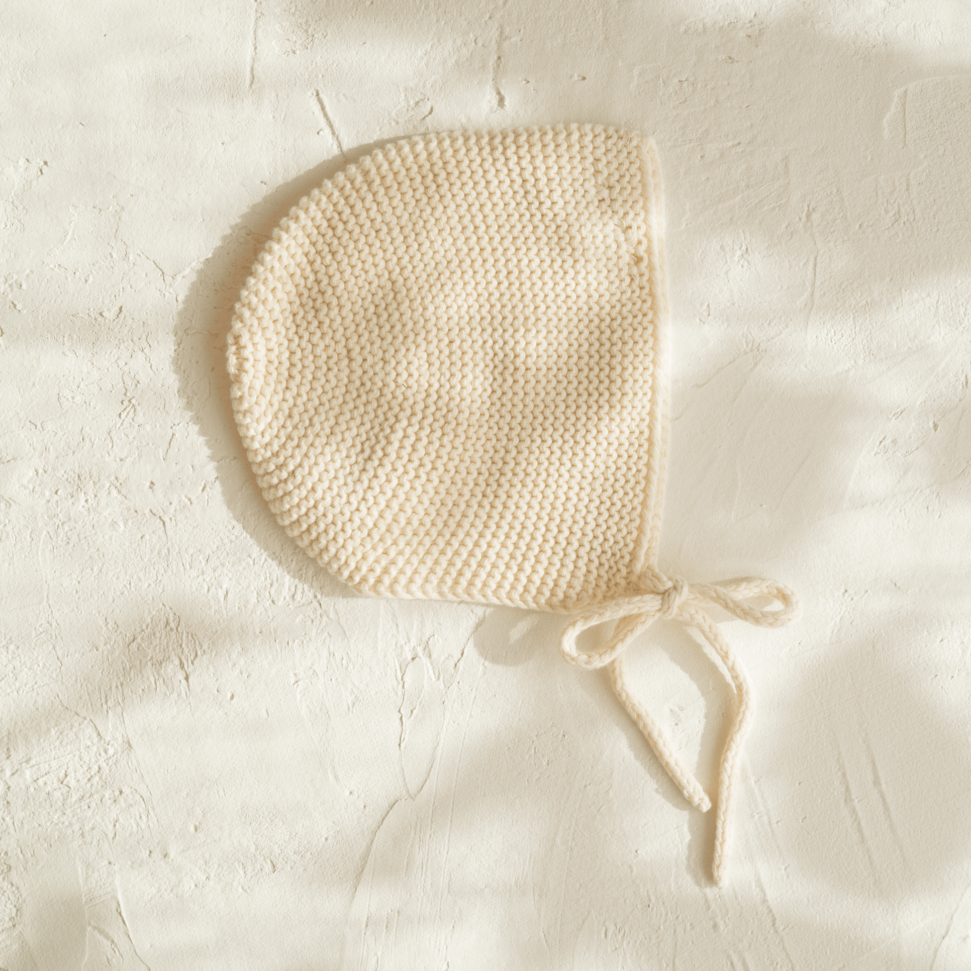 An illoura baby bonnet | vanilla on a white wall.