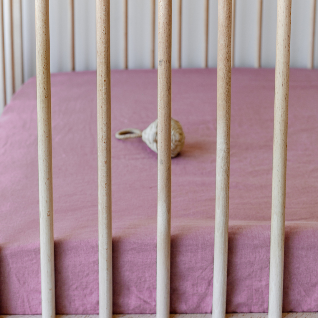 A baby crib with a Warren Hill french linen fitted cot sheet | smoke pink on it.