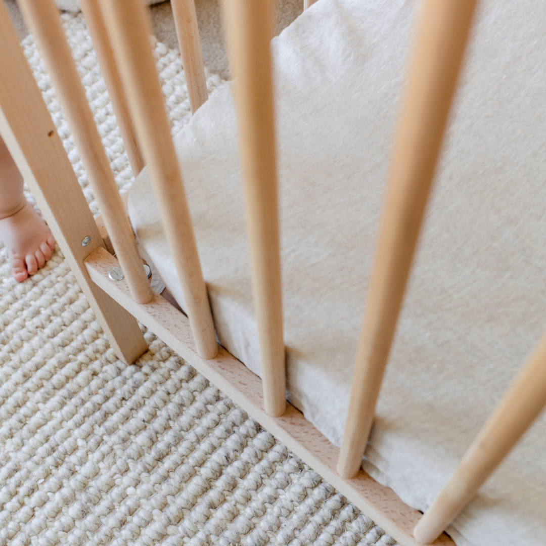 A baby's feet standing on a Warren Hill french linen fitted cot sheet in a crib.