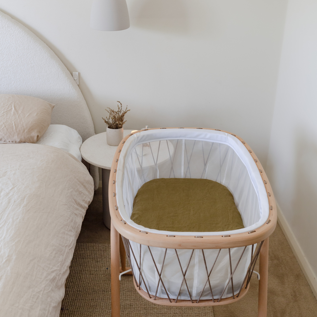 A neutral nursery with a baby's cot, a bed, and a bedside table adorned with Warren Hill french linen fitted bassinet sheets in olive.