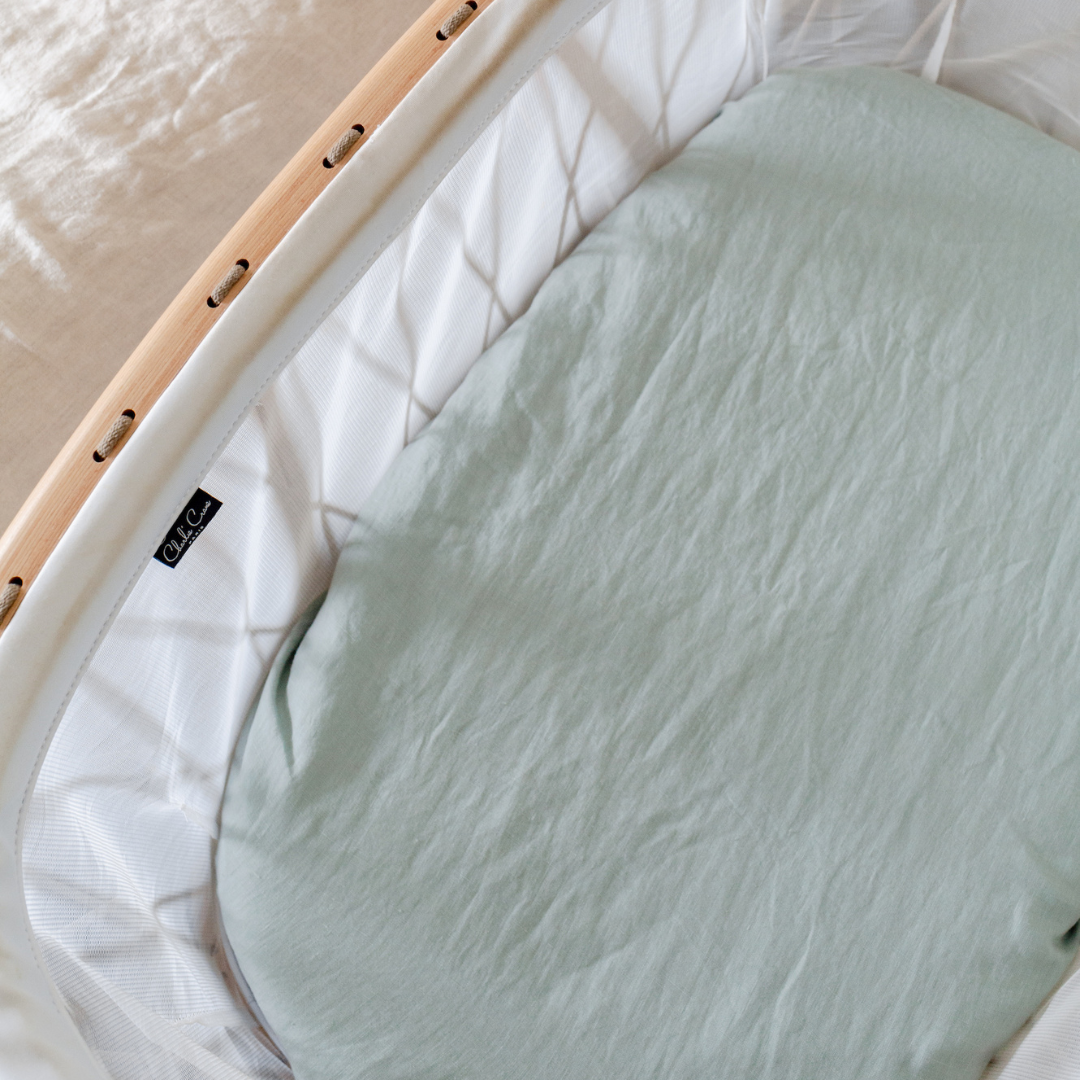 A neutral nursery with a Warren Hill french linen fitted bassinet sheet in aqua on a baby's cot.