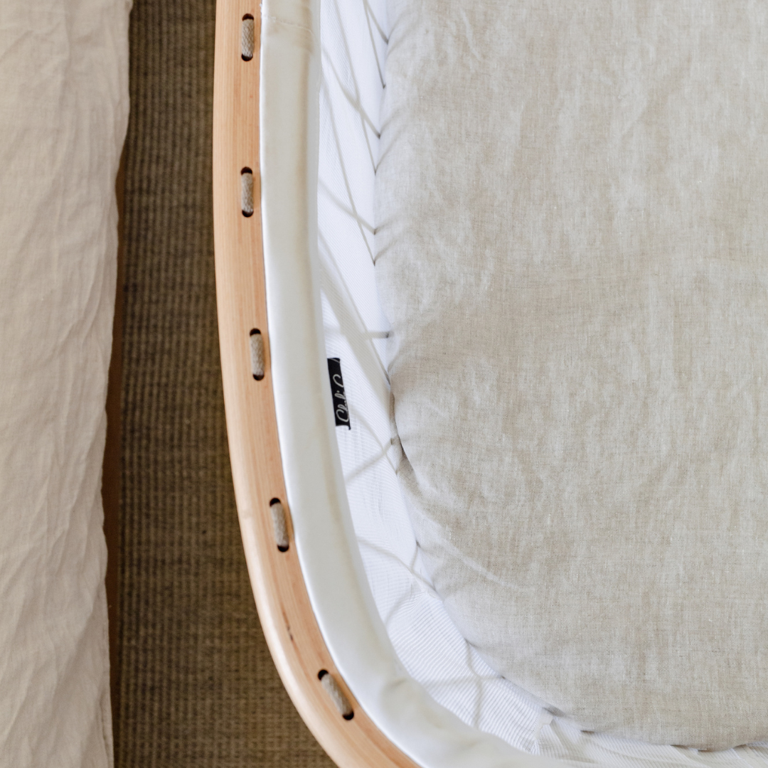 A close up of a bed with Warren Hill french linen fitted bassinet sheet in natural and a wooden frame.