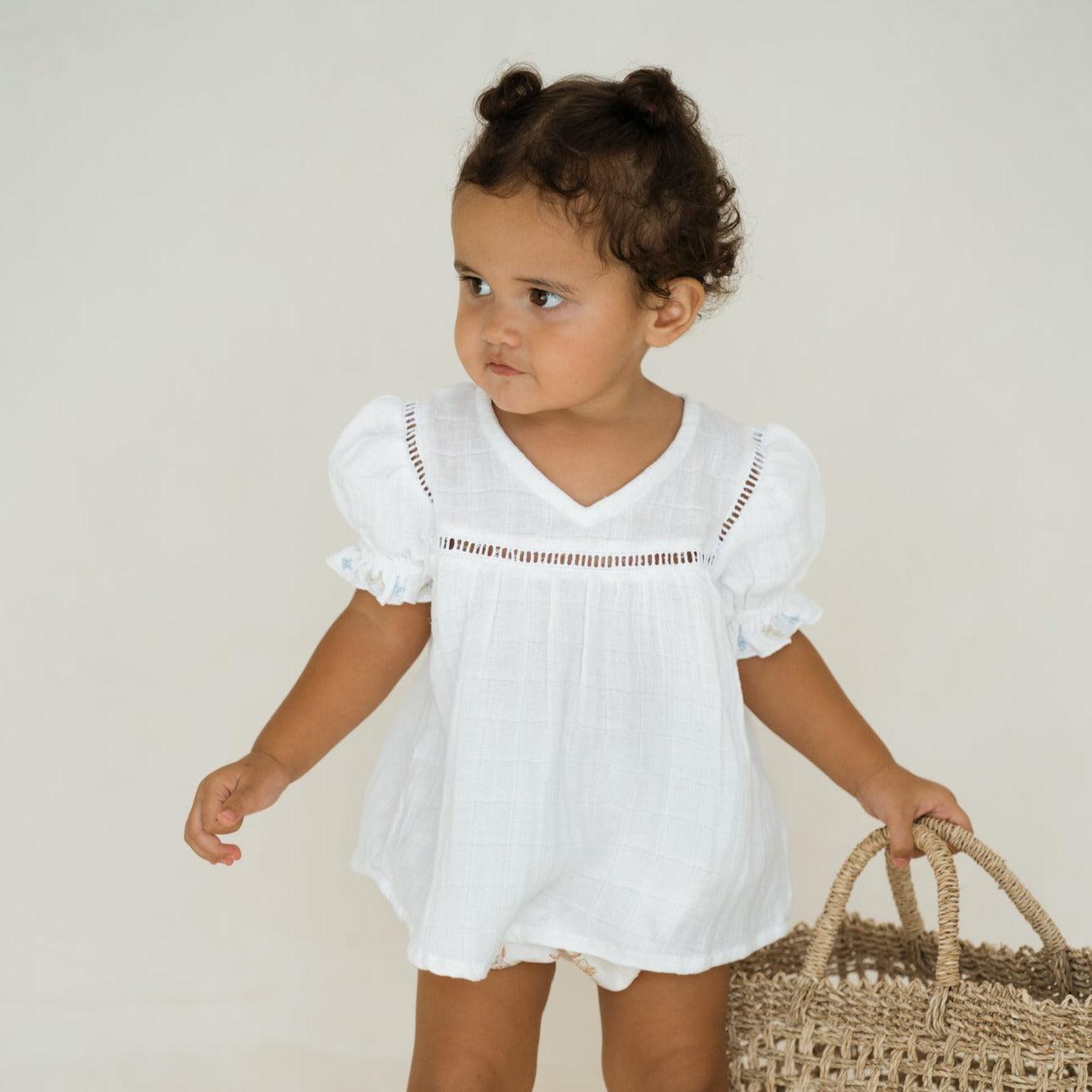 A baby girl wearing Illoura the Label's hand-embroidered floral-trimmed white clover blouse & bloomer set and carrying a vintage-inspired basket.