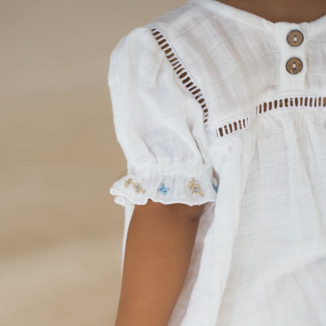 An Illoura the Label clover blouse & bloomer set with hand-embroidered floral trim worn by a little girl in a white dress.