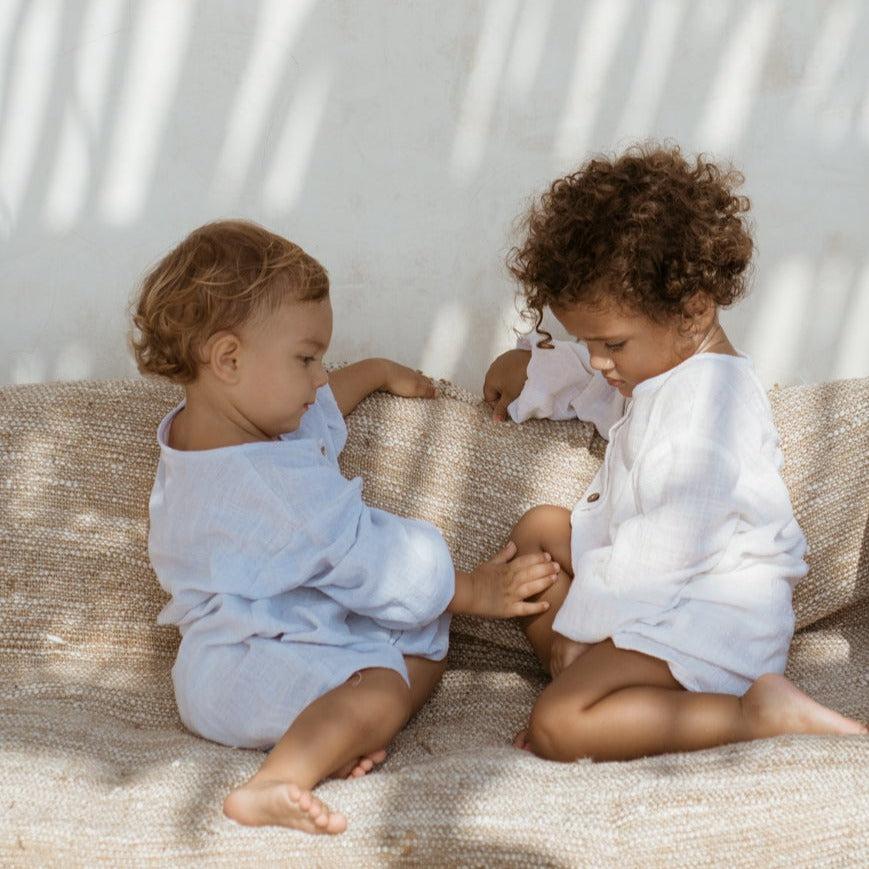 Two young children sitting on a couch wearing Illoura the Label's husk romper in powder blue.
