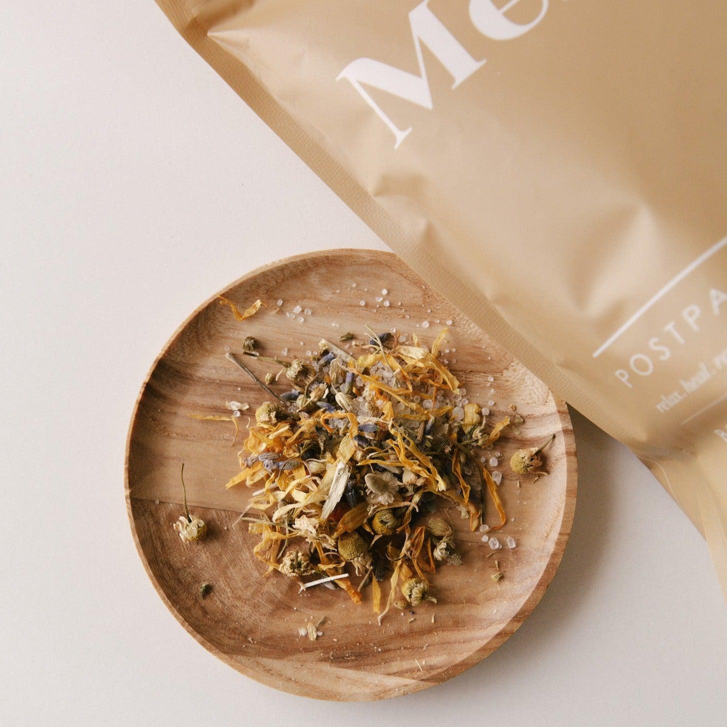 A wooden plate with dried herbs next to a bag of tea featuring Mère postpartum bath salts.