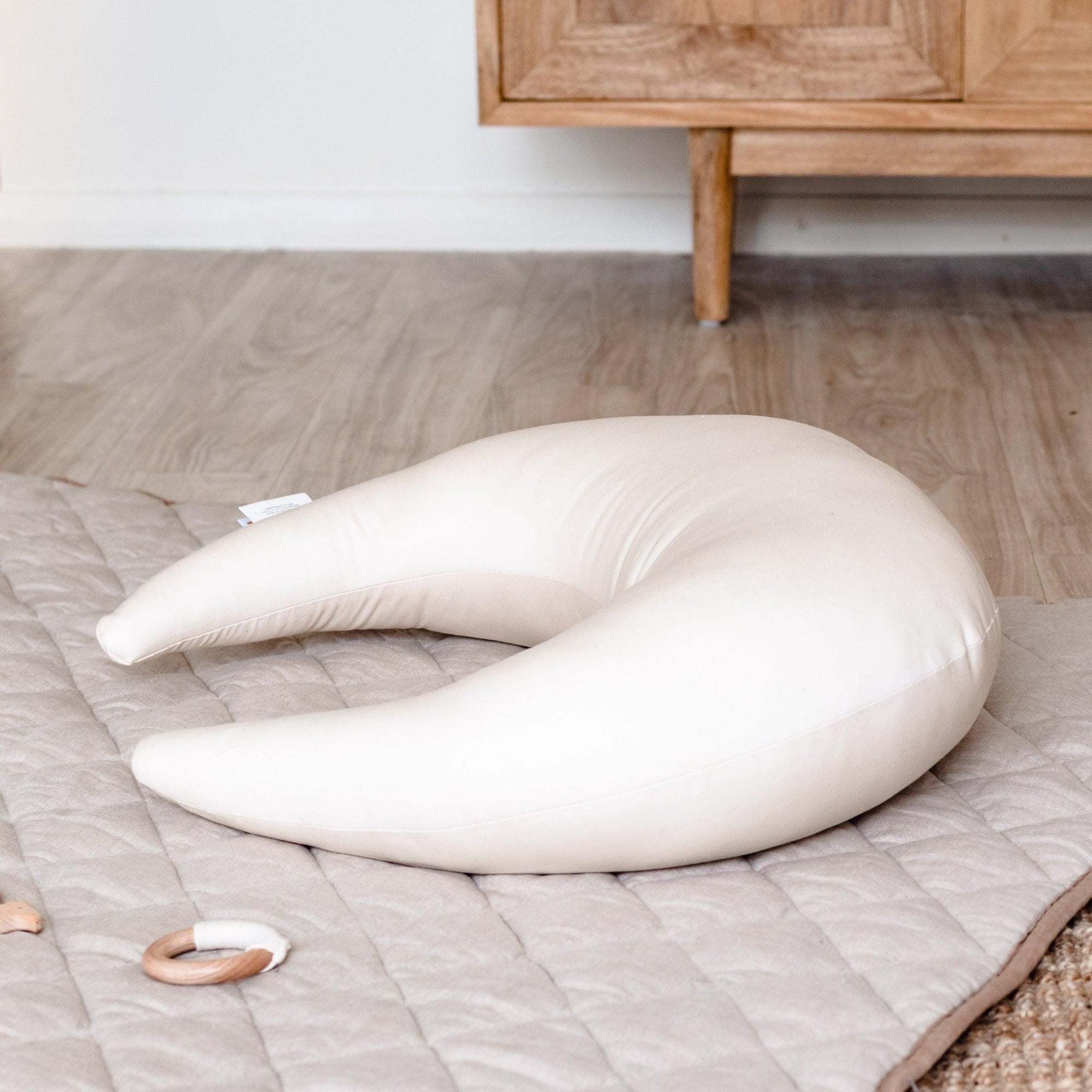 A white Snuggle Me Feeding & Support pillow on a rug next to a wooden toy.