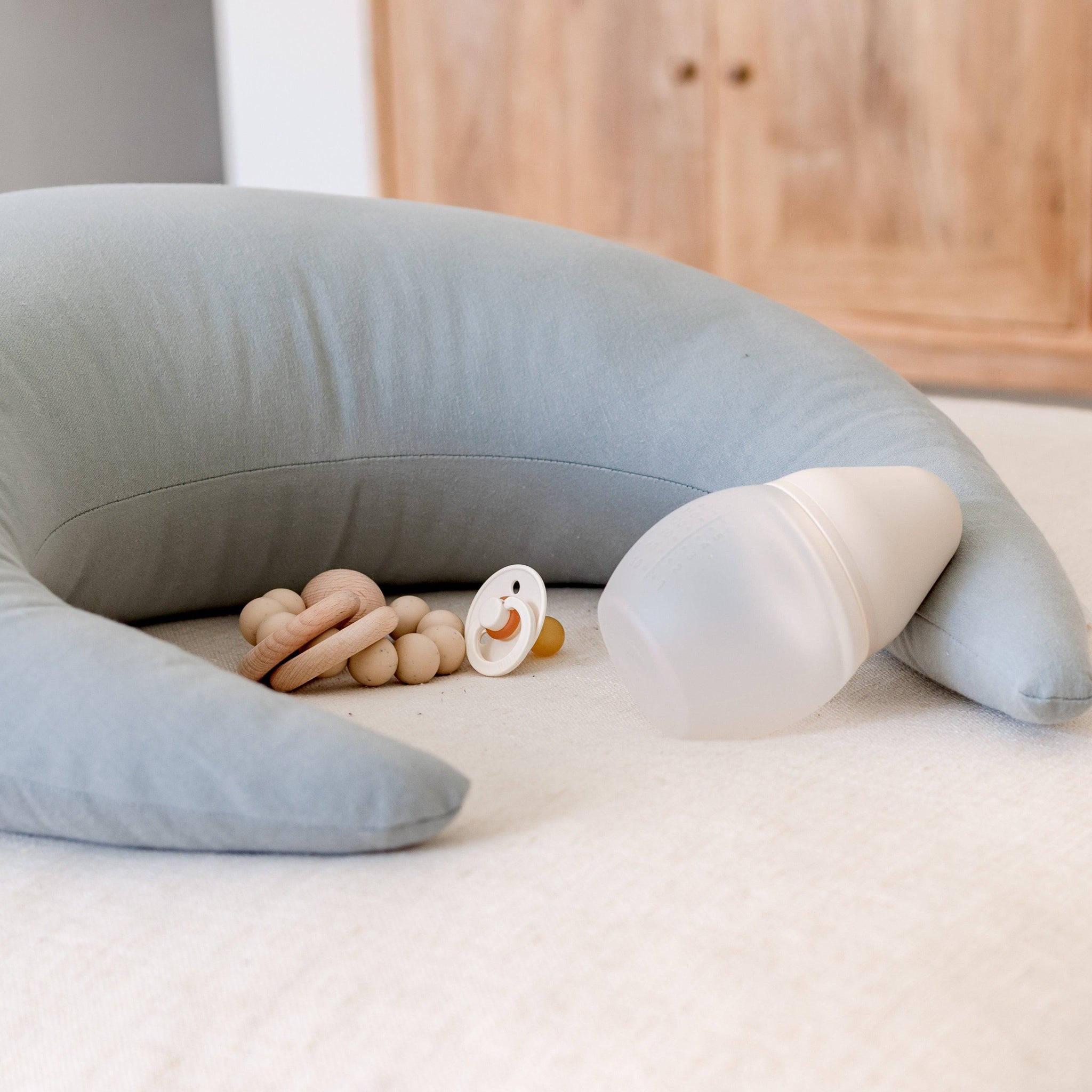 A Snuggle Me Feeding & Support Pillow in a crescent shape in the shade Slate on a bed next to a bottle and dummy.